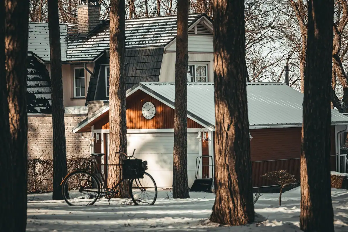 Casă de lemn iarna, cu bicicletă sprijinită în față, între pini înzăpeziți