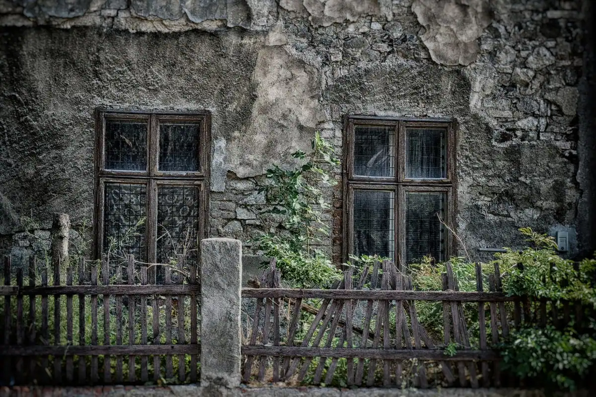 Casă veche din piatră cu două ferestre și un gard de lemn în față