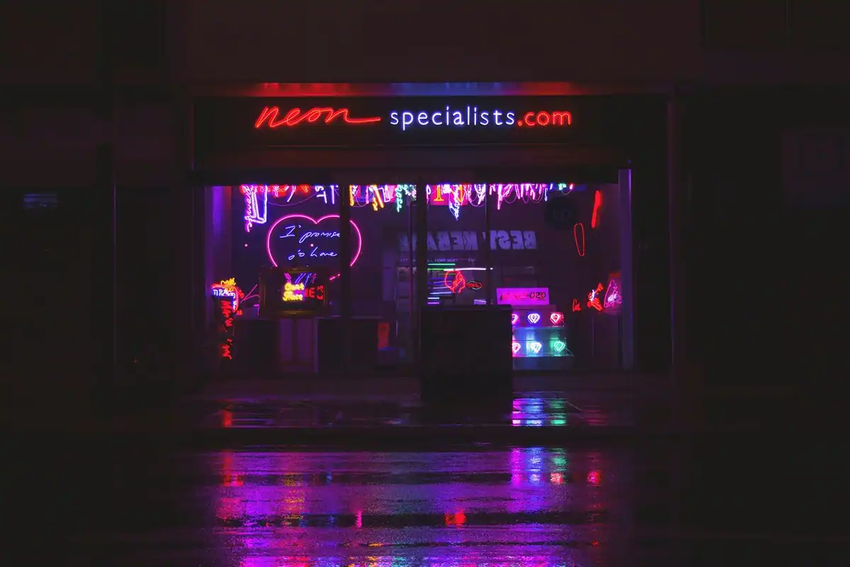 Vitrină de magazin pe o stradă noaptea, cu reclame cu neon aprinse reflectate pe asfaltul ud