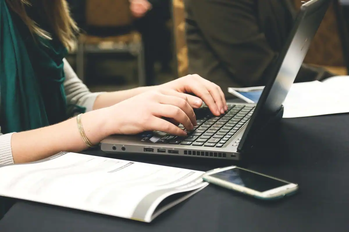 Femeie care scrie pe laptop, lângă un document și un telefon